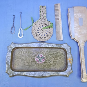 May include: An assortment of vintage vanity items on a blue surface. Includes a crochet bag, comb, hand mirror, and a decorative tray with a floral design. Also present are two metal tools, possibly for crochet or embroidery.
