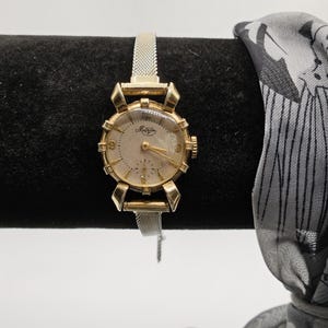 May include: A gold-toned wristwatch with a white face and black Roman numeral markings. The watch has a mesh metal band and the words "Mathey Tissot" are printed on the face.
