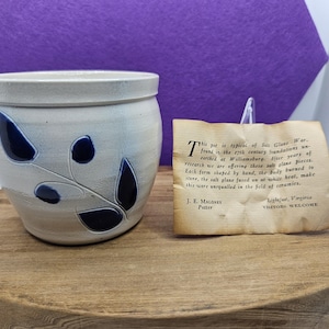 May include: A ceramic pot with a cream-colored base and dark blue leaf design. The pot is on a wooden surface, with a handwritten note beside it. The note describes the pot's origin and maker.
