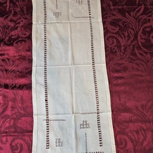 May include: Ivory-colored rectangular table runner with embroidered floral and geometric patterns. The runner features a decorative border with small, evenly spaced cut-out details. The design includes floral and geometric shapes.