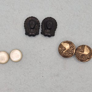 May include: An assortment of vintage buttons and embellishments. Two dark brown, detailed face-shaped buttons, two gold-colored, ornate round buttons, and two small, round, off-white buttons are displayed on a light gray surface.