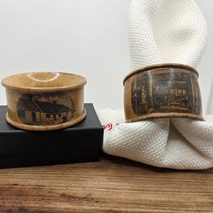May include: Two wooden napkin rings with detailed black and white illustrations. One ring features a house, the other a church interior. The rings are on a wooden surface, one on a black box, and the other on a white napkin.
