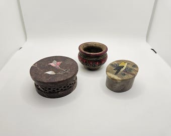 May include: Three decorative stone containers. One is a small, round, dark brown box with a carved floral design. Another is a small, round, red and brown pot. The third is a small, round box with a bird design.