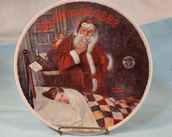 May include: A white ceramic plate with a Christmas-themed illustration. The image depicts Santa Claus looking over a sleeping child in bed. The text "Christmas 1986" is printed in red on the plate. The artist's name, "Norman Rockwell", is printed in red at the bottom of the plate.