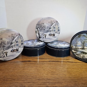 May include: A collection of vintage round tins with winter landscape scenes. The tins feature illustrations of houses, trees, and people in a snowy setting. The tins are various sizes and have dark blue sides.