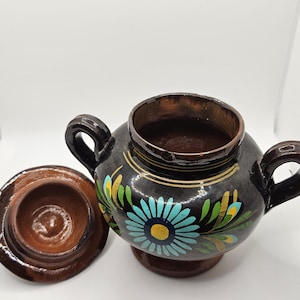 May include: A handcrafted, dark brown ceramic sugar bowl with a lid, featuring a vibrant floral design in blue, yellow, and green. The bowl has two handles and a pedestal base. The lid is off to the side.
