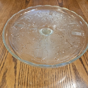 May include: Clear glass cake stand with a scalloped edge and embossed floral and geometric patterns. The stand has a central raised area for stability and is designed to display cakes and other baked goods.