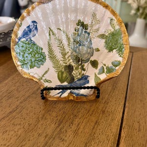 Puede incluir: Un plato decorativo en forma de concha con un borde dorado. El interior blanco presenta un diseño floral y de pájaros en azul y verde. El plato está sostenido por un soporte de metal negro, mostrando una estética única inspirada en la naturaleza.