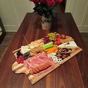 May include: A wooden charcuterie board with a variety of snacks, including crackers, cheese, salami, pretzels, nuts, and fresh fruit. A bouquet of red roses in a ceramic vase sits on a dark wood table.