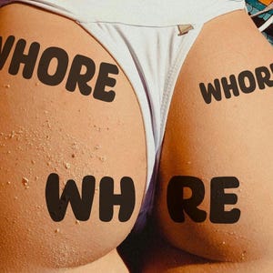 May include: Close-up of a person's buttocks with the word "WHORE" printed on the skin in black, bold letters. The person is wearing a white bikini bottom. Sand-like particles are visible on the skin.