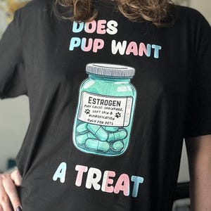 May include: Black t-shirt with the text "Does pup want a treat" in pink and blue. A cartoon illustration of a pill bottle labelled "Estrogen" with blue pills inside.