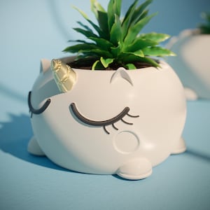 May include: White ceramic planter shaped like a unicorn with closed eyes and a gold horn. The planter is filled with a green succulent plant.