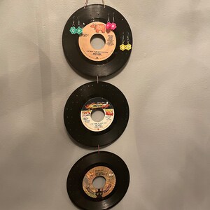 Puede incluir: Decoración de pared hecha con tres discos de vinilo negros vintage, cada uno con una etiqueta diferente. El disco superior tiene tres pares de pendientes de flores. Los discos están suspendidos por una cuerda.