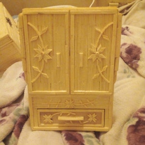 Puede incluir: Un pequeño gabinete de madera con dos puertas y un cajón. El gabinete está hecho de cerillas y tiene un diseño floral en la parte delantera.