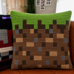 May include: A square crochet pillow featuring a pixelated design of a grass block. The top section is bright green, while the lower portion consists of a grid of brown, tan, and grey squares, mimicking a dirt block texture with a visible yarn stitch.
