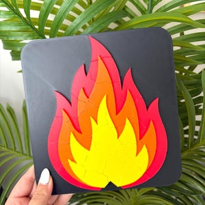 May include: A square black plaque featuring a layered flame design composed of interlocking pieces in vibrant shades of red, orange, and yellow. The flame graphic is centered on the dark base, creating a high-contrast visual effect with distinct color segments.