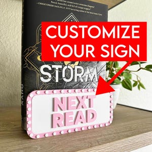 May include: A pink and white sign with the text "NEXT READ" in front of a book. The sign is rectangular with a pink border and white dots. The book has the word "STORM" on the cover. The image also includes the text "CUSTOMIZE YOUR SIGN".
