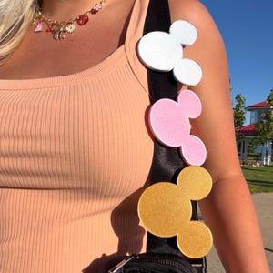 May include: A black bag strap adorned with three circular mouse-ear shaped attachments in white, pink, and gold glitter finishes. The strap is worn over a peach-colored ribbed tank top, accompanied by a gold chain necklace with various small charms.