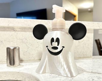 May include: A white and black soap dispenser shaped like a ghost with Mickey Mouse ears. The dispenser has a smiling face cut out of the front and a pump on top. A stainless steel tumbler is to the left of the dispenser.