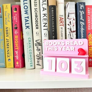 May include: A pink and white book counter displaying "BOOKS READ THIS YEAR" and the number "103". A small red heart is on the side. The counter is in front of a shelf of books.