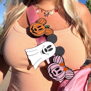 May include: A pink crossbody bag strap adorned with three character-themed patches: an orange pumpkin with mouse ears, a white ghost with black mouse ears, and a light pink pumpkin with a bow and mouse ears, worn over a peach-colored ribbed tank top.
