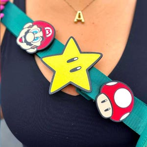 May include: A teal green strap adorned with colorful cartoon character pins. The pins include a yellow star, a red mushroom, and a Mario character. The strap is worn across the body.