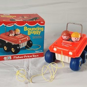 Puede incluir: Un juguete Fisher-Price Bouncing Buggy rojo y azul con una cuerda para tirar. El juguete tiene dos figuras en el asiento delantero y una caja blanca en la parte trasera. La caja tiene el texto "Bouncing Buggy" en ella. El juguete está sobre una superficie blanca.