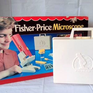Puede incluir: Un conjunto de juguete vintage de microscopio Fisher-Price en una maleta de transporte de plástico blanco. La caja muestra a un niño usando el microscopio. La maleta tiene el logotipo de Fisher-Price.