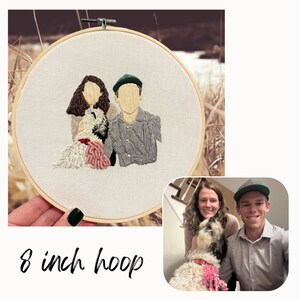 Op de afbeelding: Borduurwerk van een portret van een stel en hun hond in een hoepel van 20 cm. Het stel is in een eenvoudige stijl afgebeeld, de vrouw links en de man rechts. De hond zit in het midden tussen hen in. Het borduurwerk is gemaakt op witte stof.