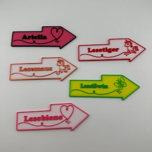 Könnte beinhalten: Eine Sammlung farbenfroher, pfeilförmiger Namensschilder. Jedes Schild zeigt einen anderen Namen oder ein Wort in einer verspielten Schriftart, zusammen mit dekorativen Elementen wie Herzen, Schmetterlingen und einem Einhorn. Die Schilder sind in Rosa-, Orange-, Rot- und Grüntönen gehalten.