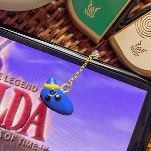 May include: A blue Ocarina of Time charm with a gold chain hanging from a Nintendo Switch console playing The Legend of Zelda: Ocarina of Time. The game screen shows Link riding Epona on a dirt path.