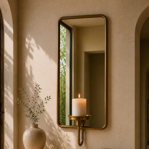 May include: A beige stone console table with two cylindrical legs, a rounded rectangular mirror with a brass frame, a wall-mounted brass candle holder with a white pillar candle, a beige ceramic vase with green sprigs, and a beige bowl.