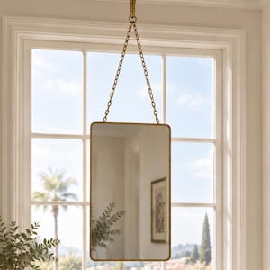 May include: A rectangular wall mirror with a thin metallic gold frame, suspended from the ceiling by two gold chains attached to a circular gold mount. The mirror features rounded corners and reflects the interior space and a window view.