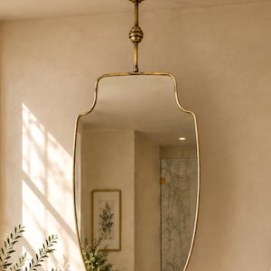 May include: A wall-mounted mirror with a brass-toned metal frame featuring a unique, elongated, curved shape. The mirror is suspended from the ceiling by a matching brass-toned metal rod with a decorative spherical accent piece.