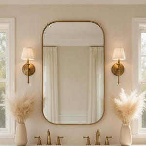 Puede incluir: Un mueble de baño con un espejo grande con marco dorado y dos apliques de pared. El mueble tiene una encimera de mármol, dos lavabos y armarios beige. Dos jarrones con hierba de la pampa seca están sobre la encimera.