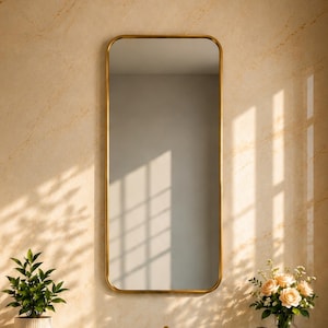 Puede incluir: Un espejo de pared rectangular con esquinas redondeadas y un marco dorado fino. El espejo está montado verticalmente sobre una encimera de mármol verde con un lavabo, grifos dorados, una planta en maceta, toallas blancas y flores.
