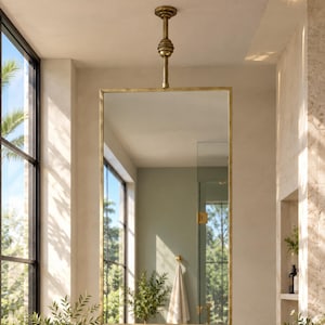 May include: A rectangular wall mirror with a thin brass frame, suspended from the ceiling by a matching brass rod. The mirror is positioned above a marble countertop with a white sink and brass faucet fixtures in a bright, sunlit bathroom.