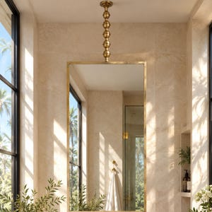Peut inclure: Un miroir mural rectangulaire avec un cadre doré fin suspendu à une tige verticale en laiton. Le miroir est placé au-dessus d'un comptoir de vanité en pierre blanche avec une robinetterie en laiton et un meuble en bois clair.