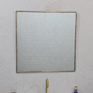 May include: A gold framed square mirror hangs above a white porcelain sink with a gold faucet. A small gold cup and a glass bottle with a purple liquid are on the counter.