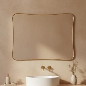 Könnte beinhalten: Ein modernes Badezimmer mit einem rechteckigen Spiegel mit goldenem Rahmen. Der Waschtisch hat ein weißes, rundes Waschbecken, goldene Armaturen und einen Holzschrank. Eine kleine Vase mit Trockenblumen und ein Seifenspender stehen auf der Theke.