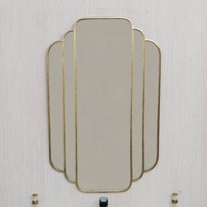 Peut inclure: Miroir de style Art Déco avec un cadre doré. Le miroir a une forme unique avec plusieurs panneaux. Un robinet noir et un évier blanc se trouvent en dessous. Deux flacons décoratifs en verre avec des accents dorés encadrent l'évier.