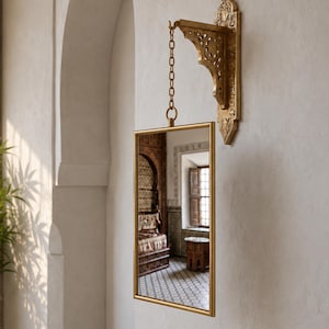 May include: A rectangular wall mirror with a thin gold frame, suspended by a metal chain from a decorative, ornate gold wall bracket featuring intricate cut-out patterns and a pointed, shield-like base mounted against a plain wall.
