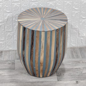 Könnte beinhalten: Ein runder Holzhocker mit einem Streifenmuster in Braun- und Blautönen. Der Hocker hat ein einzigartiges Sonnenstrahlendesign auf der Oberseite.