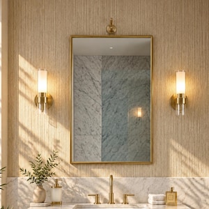 May include: A rectangular wall mirror with a thin gold frame, positioned above a floating vanity with a white marble countertop and a gold faucet. Two cylindrical wall sconces with frosted glass shades flank the mirror on a textured beige wall.