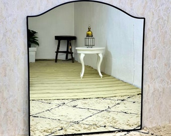 Op de afbeelding: Een zwarte, gebogen spiegel met een gefacetteerde rand, die een kamer met een witte tafel en een kruk weerspiegelt. De spiegel staat op een wit en zwart gestreept tapijt.