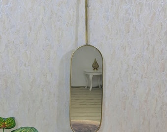 May include: A gold-toned oval mirror with a long, thin frame hangs from a ceiling mount. The mirror is reflecting a room with a white table and a decorative object on top.