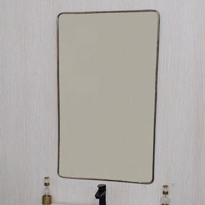 May include: A rectangular mirror with a thin, bronze-colored frame hangs above a white sink. A black faucet and two decorative bottles with gold accents are on the countertop. The wall behind the mirror has a light wood grain pattern.