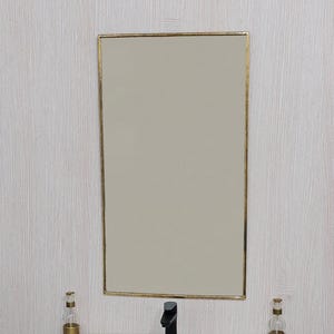 May include: A rectangular mirror with a gold frame hangs above a white sink with a black tap. Two decorative glass bottles with gold bases sit on the countertop. The wall behind the mirror has textured, light-coloured wallpaper.