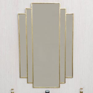 Pode incluir: Espelho em estilo Art Déco com moldura dourada, montado acima de uma pia branca. O espelho tem formato retangular com bordas escalonadas. Uma torneira preta e duas garrafas decorativas também são visíveis.