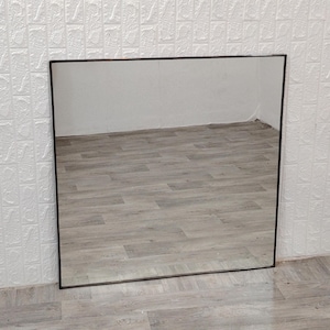 Pode incluir: Um espelho grande e quadrado com uma moldura preta fina. O espelho reflete um piso de madeira.